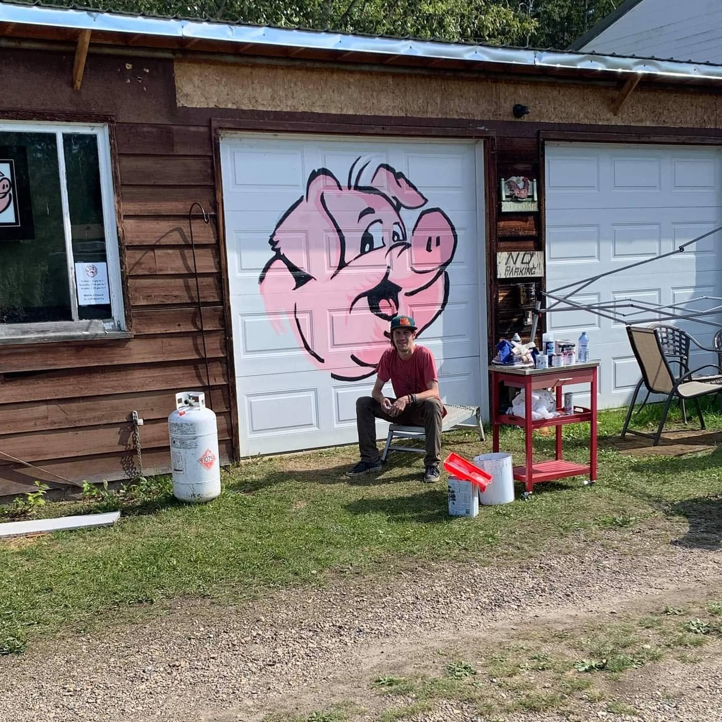 Derek Boe seated in front of the completed pink pig mural on the Hog Saloon garage door